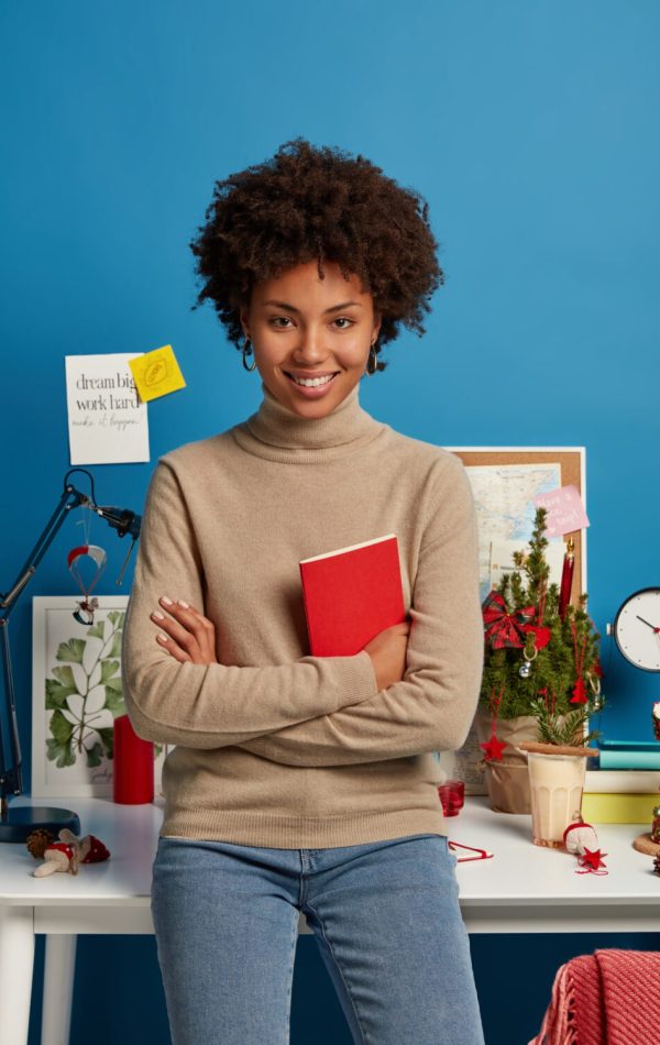 vertical-shot-pleased-curly-haired-female-teacher-prepares-lessons-home-holds-red-textbook-poses-against-desktop-e1699772972688.jpg